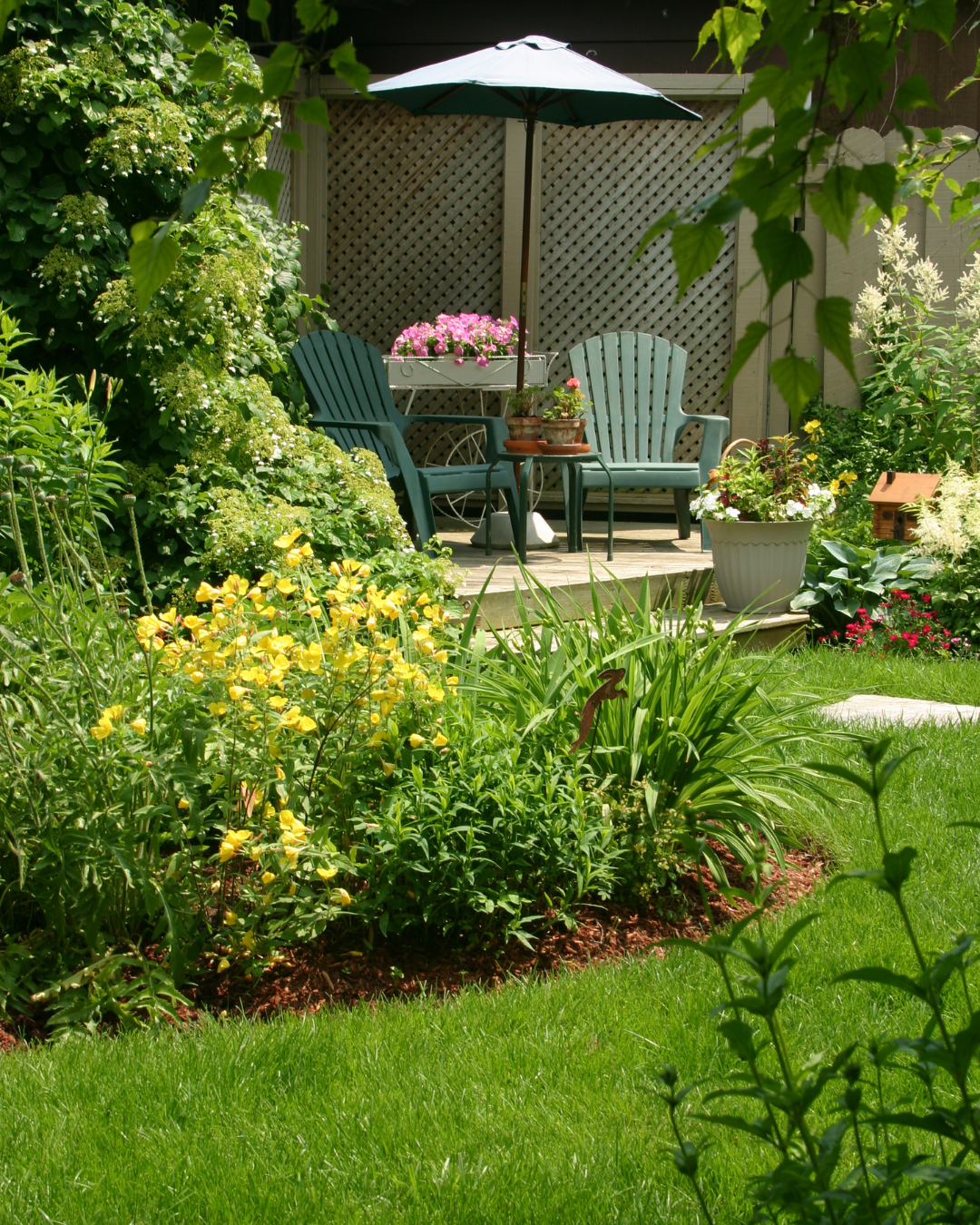 Garden seating area with two blue Adirondack chairs under a green patio umbrella beside a flower-filled table planter.